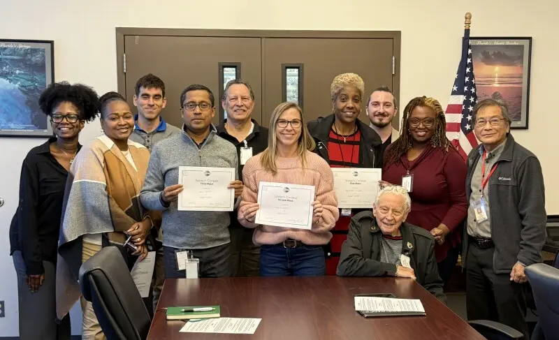 A diverse group of twelve people, some holding certificates, pose for a photo in an office-like setting. Members of the Podemos Hablar Toastmasters Club stand proudly with smiles. An American flag is in the background. Some are seated, while others stand behind them, smiling at the camera.