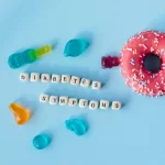 A pink donut with sprinkles sits alongside gummy candies on a blue background, where lettered cubes spell out "DIABETIC SYMPTOMS.