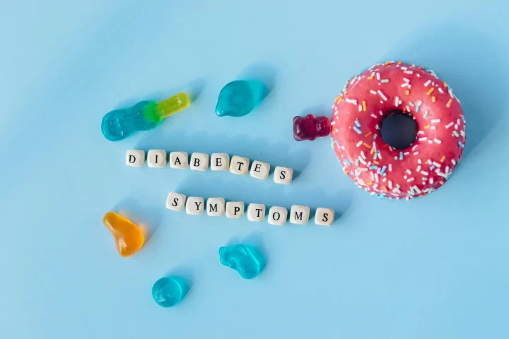 Una dona rosa con chispitas y gomitas de colores se encuentran sobre un fondo azul claro. Debajo de los caramelos, hay bloques con letras que forman la palabra "DIABETES SÍNTOMAS", lo que le da un toque divertido e informativo a la escena.