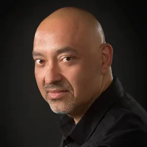 A bald man with a light mustache and goatee, possibly Humberto Castillero, is wearing a black shirt and looking intently at the camera against a dark background.