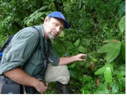 Dave Skinner, wearing a blue cap and green shirt, is carrying a backpack while crouched near a lush green plant. He is pointing his finger at the plant, inspecting it closely. Dense greenery surrounds him.