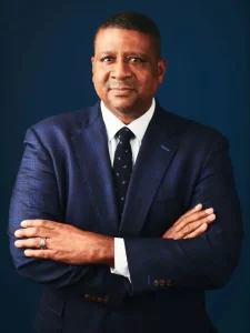 A man in a dark blue suit and tie stands confidently with his arms crossed against a dark blue background, symbolizing leadership within the minority chamber of commerce. He wears a wedding ring and looks directly at the camera.