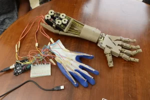 A robotic prosthetic hand rests on a wooden table, connected to an electronic circuit, showcasing the ingenuity of local students who designed a 3D prosthetic arm. Wires run from a sensor-equipped fabric glove, complete with a breadboard and microcontroller for seamless control.