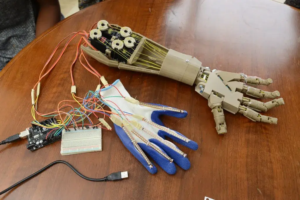 A robotic prosthetic hand rests on a wooden table, connected to an electronic circuit, showcasing the ingenuity of local students who designed a 3D prosthetic arm. Wires run from a sensor-equipped fabric glove, complete with a breadboard and microcontroller for seamless control.