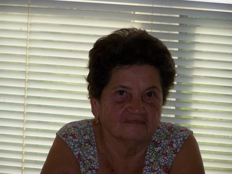 An older woman with short dark hair and a floral top sits in front of window blinds, looking at the camera with a neutral expression. The softly lit room captures the contemplative moment, hinting at fleeting thoughts of memory loss that gently weave through her mind.