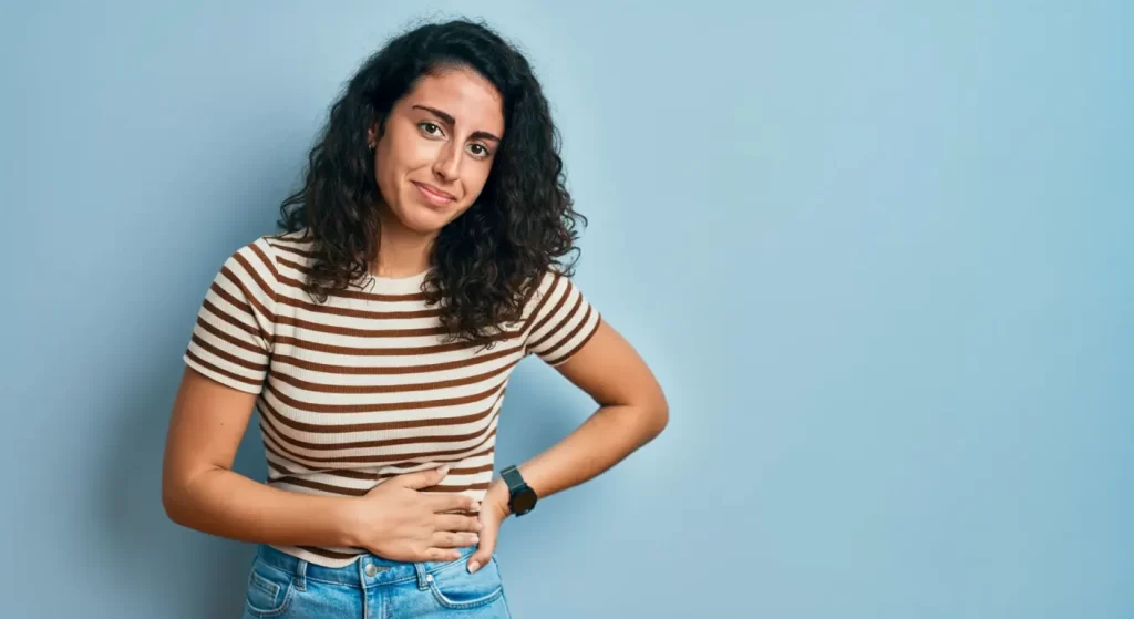 Una persona de pelo rizado, con camiseta de rayas y vaqueros, está de pie sobre un fondo azul claro. Sonríe levemente, a pesar de sentir un ligero malestar por el dolor abdominal, y tiene una mano en la cadera.