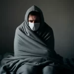 A person, seemingly battling the flu, sits on a bed wrapped in a gray blanket, wearing a matching hoodie and white face mask. The dimly lit room with its plain wall adds to the sense of solitude, reflecting the quiet battle against influenza.