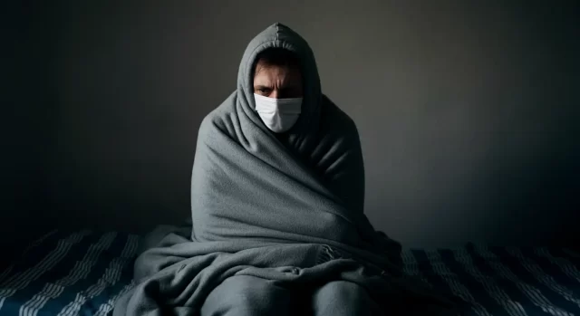 A person, seemingly battling the flu, sits on a bed wrapped in a gray blanket, wearing a matching hoodie and white face mask. The dimly lit room with its plain wall adds to the sense of solitude, reflecting the quiet battle against influenza.