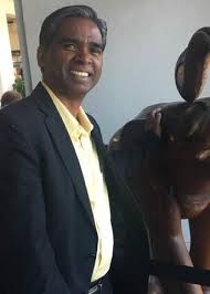 A man in a dark suit and yellow shirt is smiling while standing next to a large object, perhaps reminiscing about his volunteer work at cataract camps. The background is a neutral-colored wall.