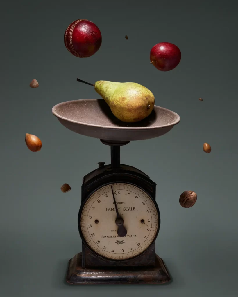 A vintage family scale holds a pear and is surrounded by levitating objects, including a cricket ball, two apples, hazelnuts, and walnuts, symbolizing a balanced diet. The scale's dial shows weight measurements in pounds and ounces against a dark green background.