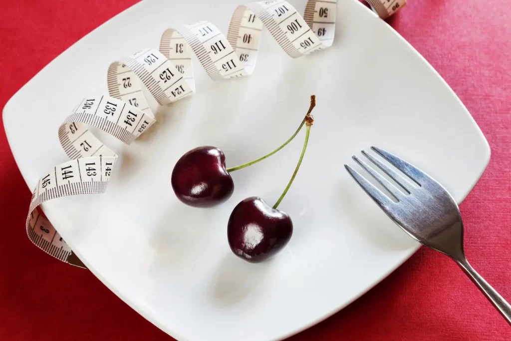 Two cherries sit on a white plate next to a fork and a coiled measuring tape, all set against a red backdrop, symbolizing weight loss and diet.