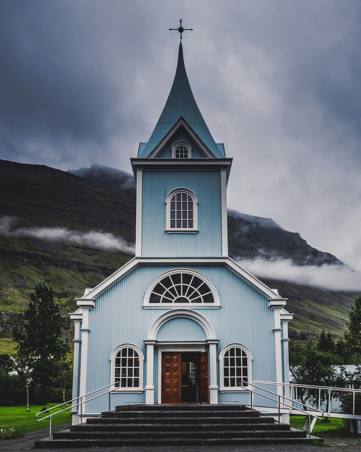 Una pequeña iglesia de color azul claro con un campanario alto coronado por una cruz se alza sobre un fondo de montañas brumosas y un cielo nublado. Su iglesia cuenta con grandes ventanales arqueados, una entrada con puerta doble de madera y una rampa que conduce a los escalones de entrada. Es verdaderamente Tu iglesia, donde la gente viene a buscar consuelo en la presencia del Señor.