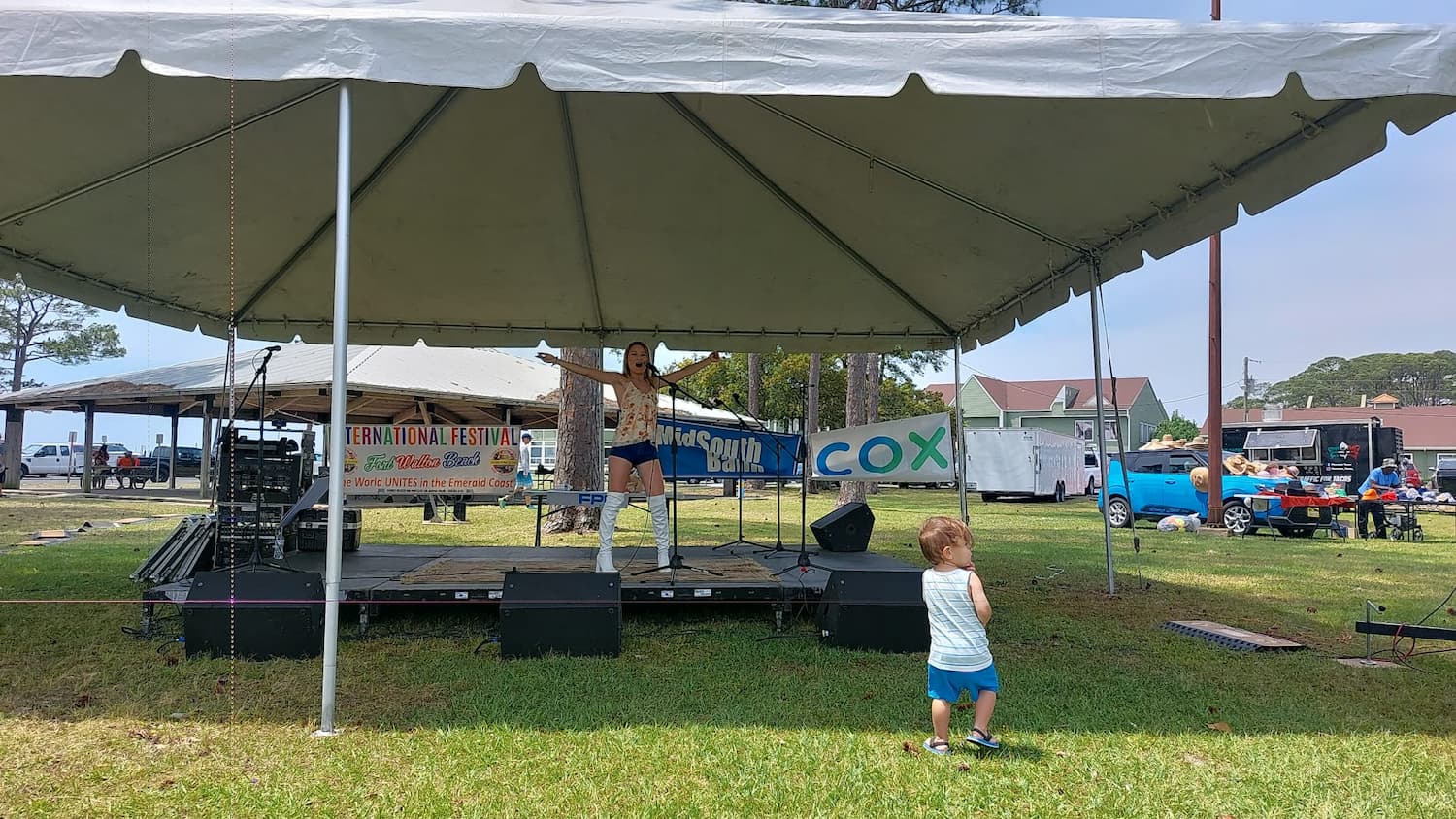 Una joven intérprete canta en un escenario al aire libre bajo una carpa blanca durante el Festival Internacional de Fort Walton Beach, con carteles que dicen "Festival Internacional" y "COX" detrás de ella. Un niño pequeño con pantalones cortos azules y una camisa blanca está de pie en el césped cerca, mirando la actuación.