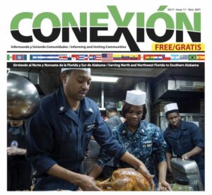 Un grupo de efectivos de la Marina, uniformados, sirve comida en la cocina de un barco. Un tripulante masculino corta un pavo cocinado mientras otros lo ayudan y observan. La portada de la edición mensual de la revista "Conexión" incluye texto multilingüe y banderas nacionales.