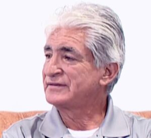 Se ve de perfil a un hombre mayor de pelo blanco que exuda una expresión amable. Víctor Torres, que lleva una camiseta polo de color gris claro, está de pie sobre un fondo blanco liso.