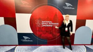 Una mujer se encuentra de pie frente a un gran telón de fondo con el texto "2024 National Small Business Conference" y los logotipos de la Asociación Nacional 8(a). Vestida de negro y con una placa con su nombre alrededor del cuello, sonríe a la cámara, encarnando el espíritu de la JNB en este importante evento.