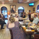 People seated at tables in a restaurant, attentively listening to a speaker at the front. Some are taking photos or videos of the event. The room is decorated with vibrant artwork.