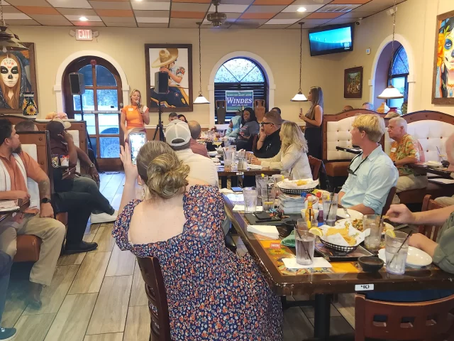 People seated at tables in a restaurant, attentively listening to a speaker at the front. Some are taking photos or videos of the event. The room is decorated with vibrant artwork.