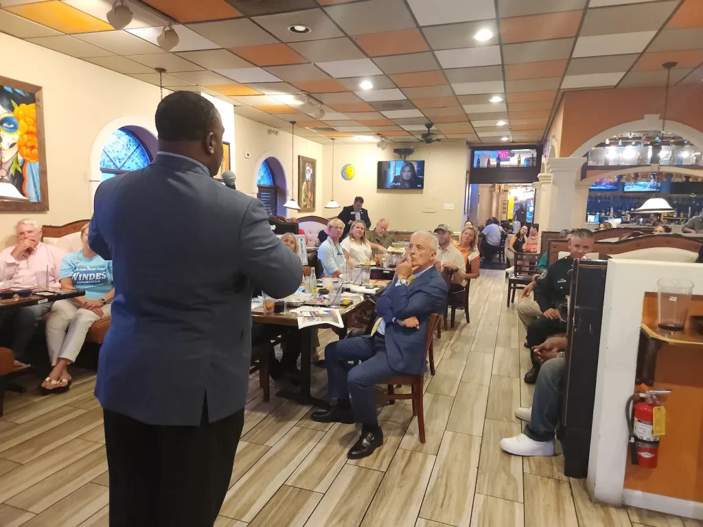 A man in a navy suit stands and speaks to a seated group of people, including several political candidates, in a restaurant with colorful decor and a tiled floor. Tables are occupied, and some people appear engaged in conversation and dining.