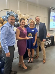 Four people standing together at an event, two men and two women, with one woman holding an award. They are smiling in front of a backdrop with balloons and a United Way banner. Casual attire suggests a celebratory occasion.