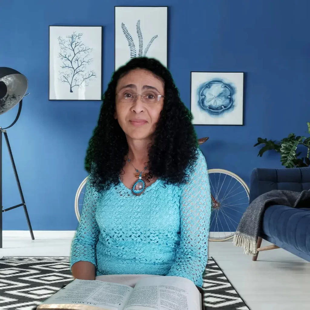 A woman with curly black hair and glasses sits in a brightly lit room with blue walls. She wears a light blue crocheted top and holds an open book on her lap. Behind her are framed artworks, a standing spotlight, a bicycle, and a blue sofa.