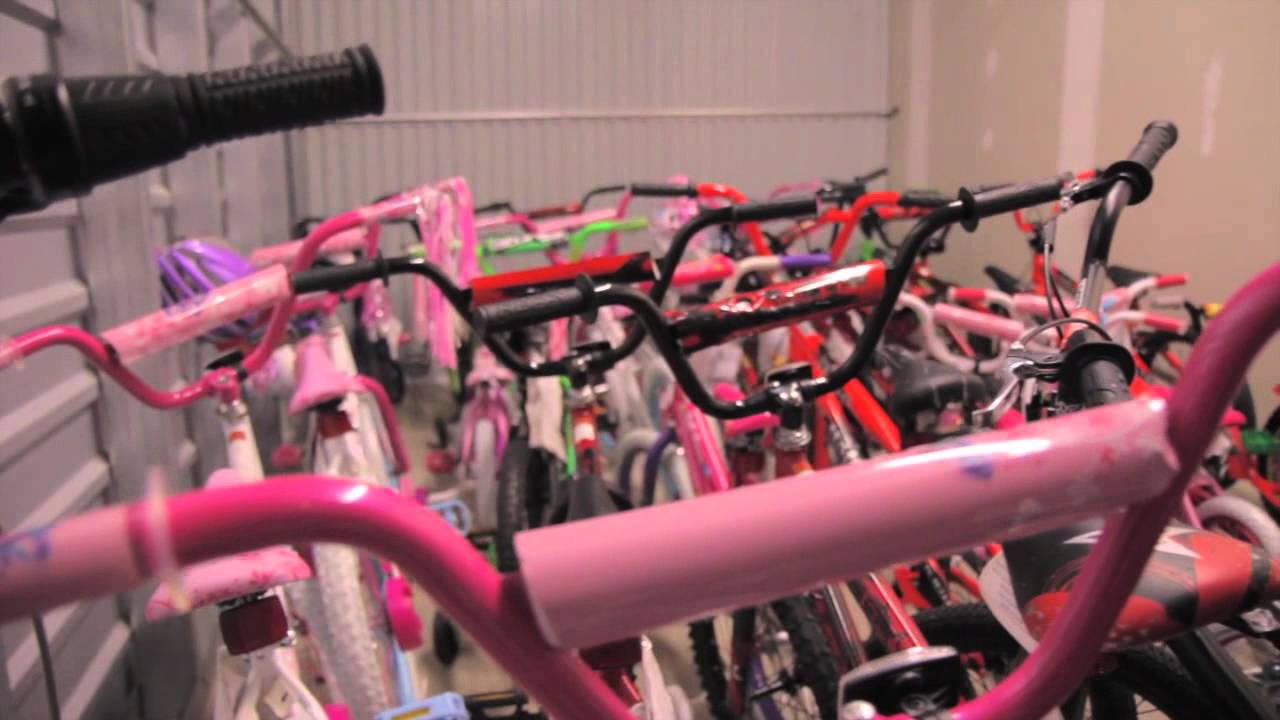 A storage room transformed into a Christmas miracle, with colorful bicycles in pink, red, and green arranged closely together. The bikes, reminiscent of a festive fiesta, feature various handlebar styles and are tightly packed within the space.