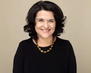 Ana Lucia Araujo, with shoulder-length dark hair, smiles warmly while wearing a black shirt and a brown beaded necklace, set against a plain beige background.