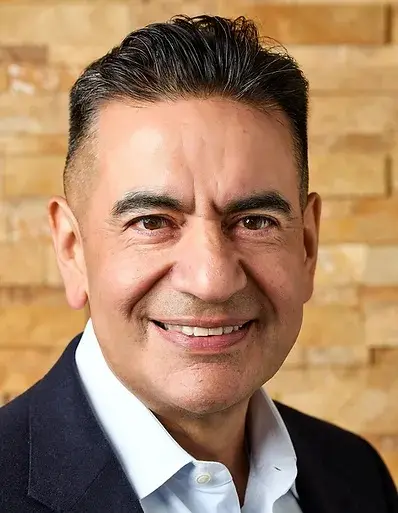 A man with short dark hair is smiling. He is wearing a white shirt and a dark blazer. The background is a tan brick wall.