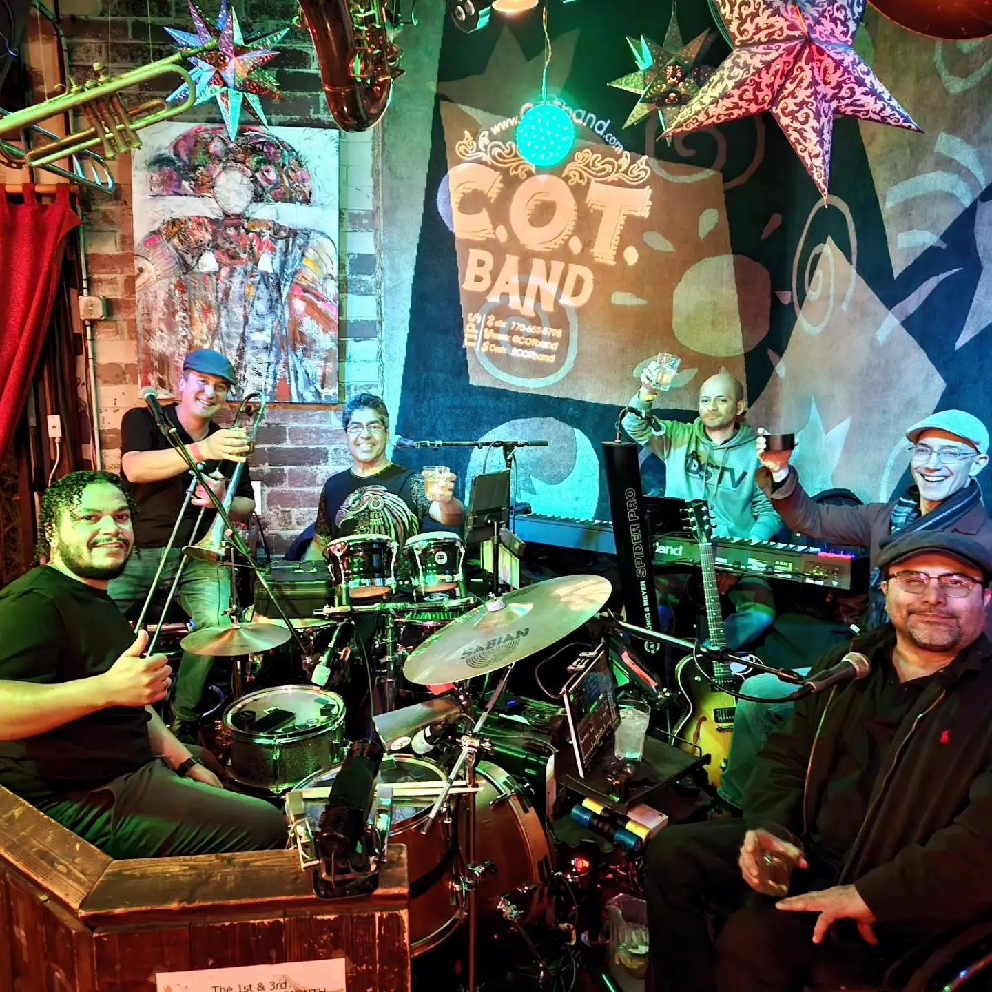 A lively band poses with instruments in a colorful, star-decorated room. Six musicians, including a drummer, saxophonist, and keyboardist, cheerfully raise their glasses. A vibrant poster in the background reads "C.O.T. Band.