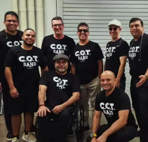 A group of eight smiling individuals, all wearing matching "C.O.T. BAND" black t-shirts, pose together indoors. One person uses a wheelchair. The background features a closed roll-up door.