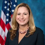 A woman with long blonde hair is smiling, wearing a dark blazer and a beaded necklace. Behind her, the American flag waves proudly against a blue backdrop, reminiscent of an auto draft caught mid-motion in the breeze.