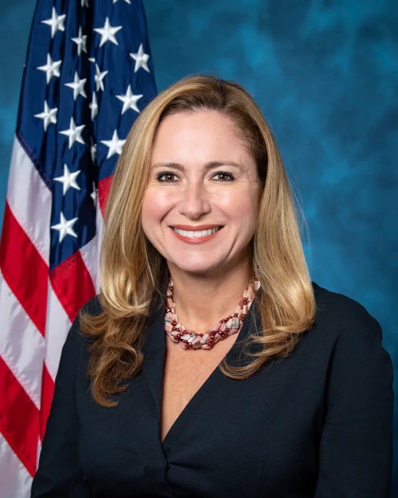 A woman with long blonde hair is smiling, wearing a dark blazer and a beaded necklace. Behind her, the American flag waves proudly against a blue backdrop, reminiscent of an auto draft caught mid-motion in the breeze.