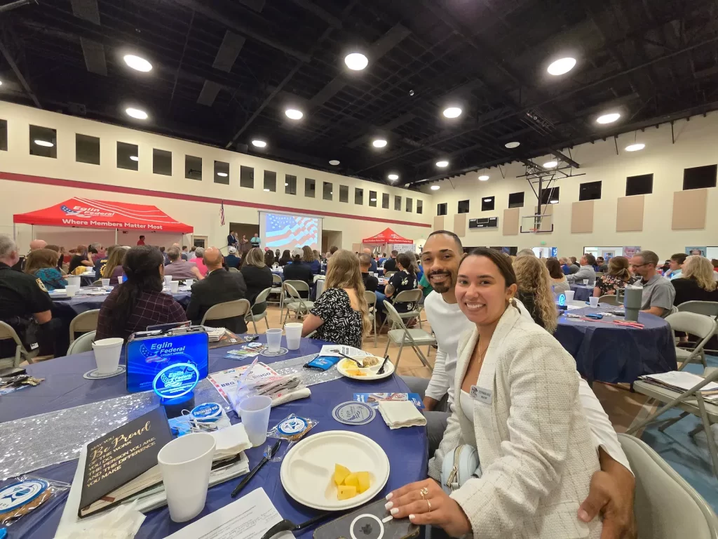 Una pareja sonriente se sienta a la mesa de un banquete en un gran pabellón deportivo decorado con pancartas y una pantalla de proyección. La sala está llena de gente sentada a las mesas, participando en un evento o reunión formal.