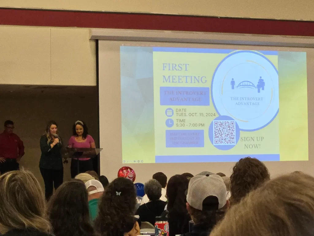 A group of people sitting in an auditorium listen to two women speaking at a podium. A large screen displays a flyer for a meeting titled "The Introvert Advantage" scheduled for October 15, 2024, from 5:30 to 7:00 PM.