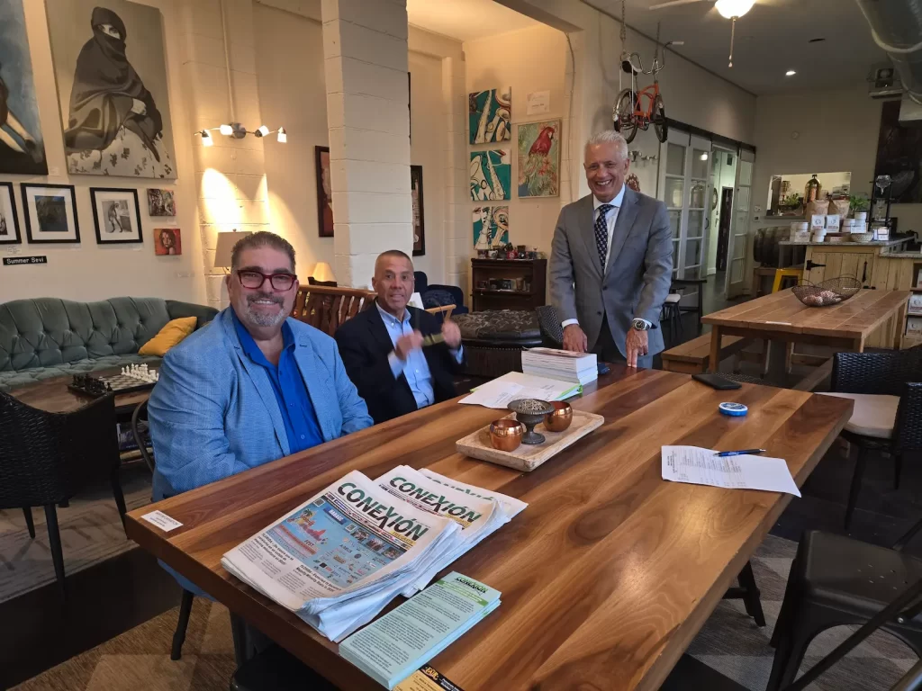 Three men in business attire are gathered around a large wooden table in a cozy, art-filled room. They are smiling and engaged in conversation. Newspapers and documents are spread across the table.