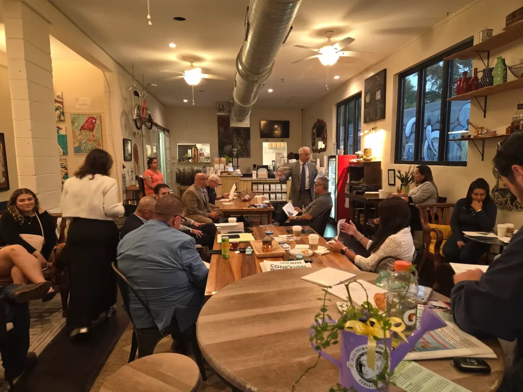 A group of people are gathered in a cozy café, some seated and others standing. There are tables with drinks and papers. The room has art on the walls and large windows. A person is speaking at the front, and everyone is engaged in the discussion.