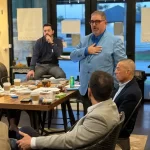 A group of men are gathered in an office setting. One man stands speaking, wearing a blue blazer, while others sit around a table with coffee cups and documents. Large windows show an outdoor view.