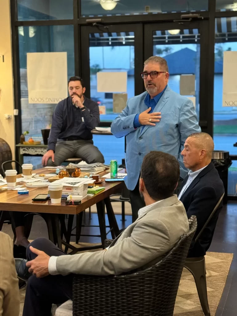 A group of men are gathered in an office setting. One man stands speaking, wearing a blue blazer, while others sit around a table with coffee cups and documents. Large windows show an outdoor view.