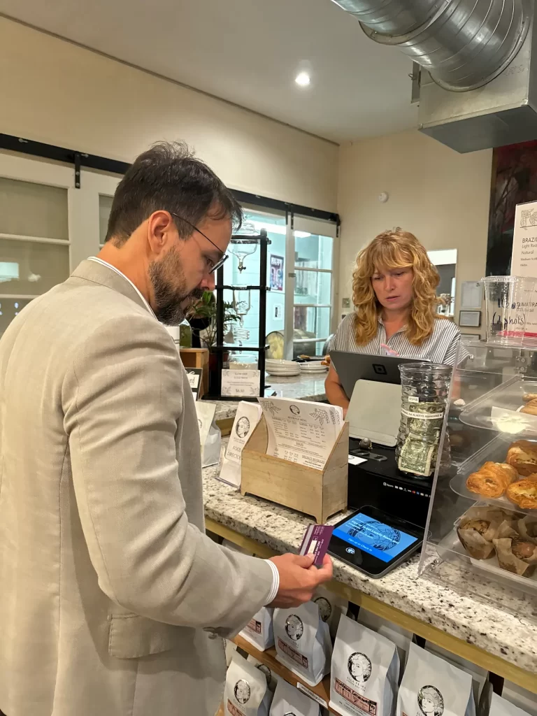 A man in a gray suit is paying with a card at a bakery counter. A woman with curly blonde hair stands behind the counter, using a touchscreen device. Pastries and a tip jar are visible on the counter.