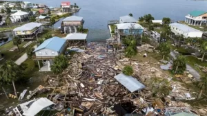 Aerial view of a coastal neighborhood devastated by Hurricane Helene reveals the impact of climate change. Debris, including broken wood and roofing, is scattered across the area. Houses show visible damage. The ocean looms in the background with several palm trees lining the area.