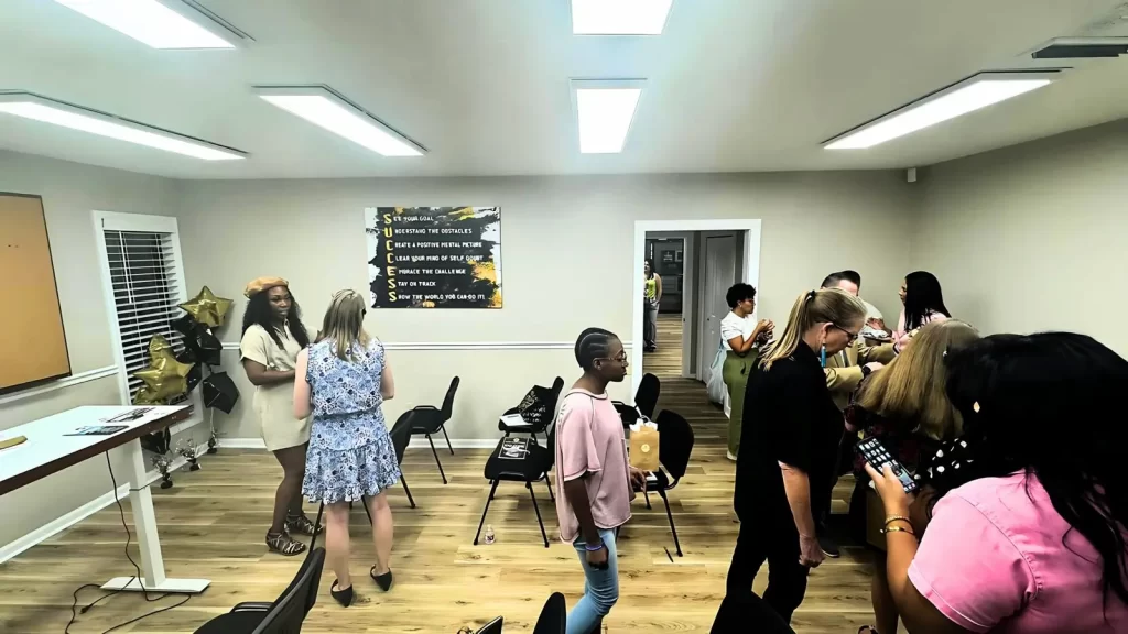 People are gathered in a brightly lit room with chairs. Some are conversing in small groups. A motivational sign is on the wall. The floor is wooden, and there are windows on the left.