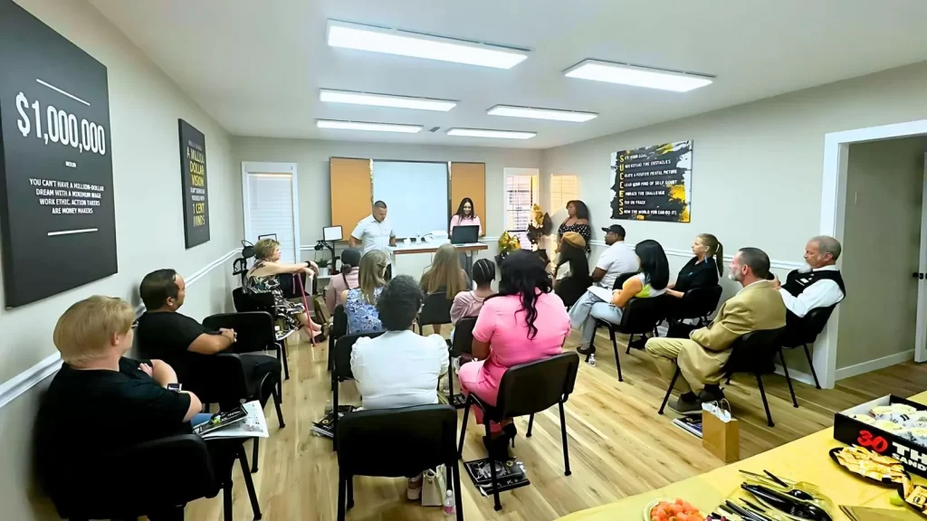 Un grupo de personas se sienta en una sala bien iluminada frente a un presentador y una pizarra. Las paredes están decoradas con carteles motivacionales. Se ven bocadillos en una mesa pequeña en primer plano.