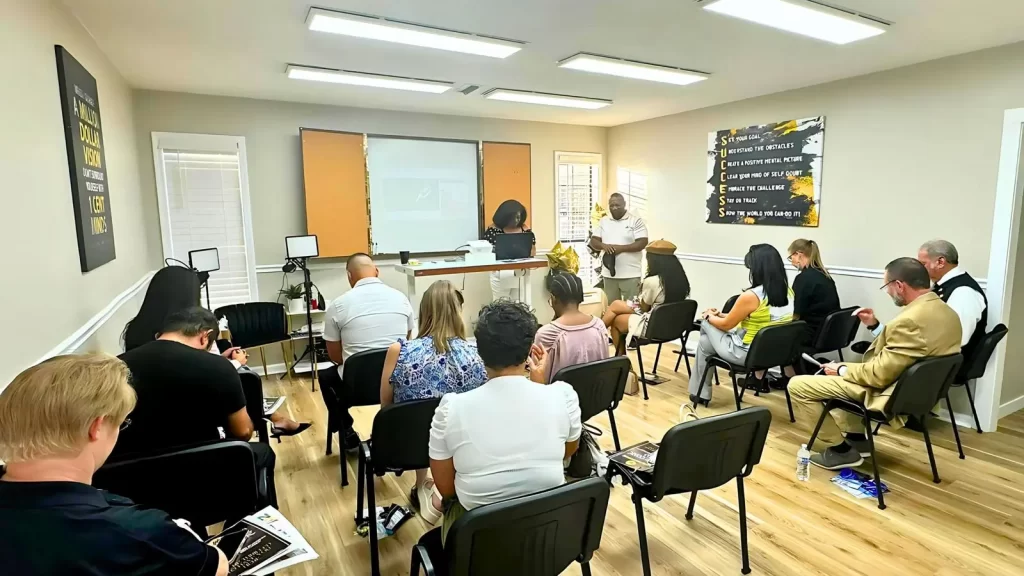 A diverse group of people seated in a small classroom facing a presenter. The presenter, standing near a whiteboard and projector screen, appears to be delivering a lecture. Attendees are taking notes and focusing on the presentation.