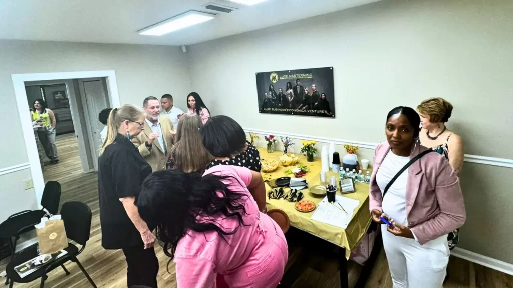 A group of people are gathered in a room with a table set with food and drinks. Some are chatting, while others are getting refreshments. A banner is on the wall. The atmosphere is casual and social.