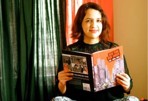 A person with shoulder-length hair is sitting indoors, smiling and holding an open book titled "New York Never Again." The room has dark curtains and soft lighting.