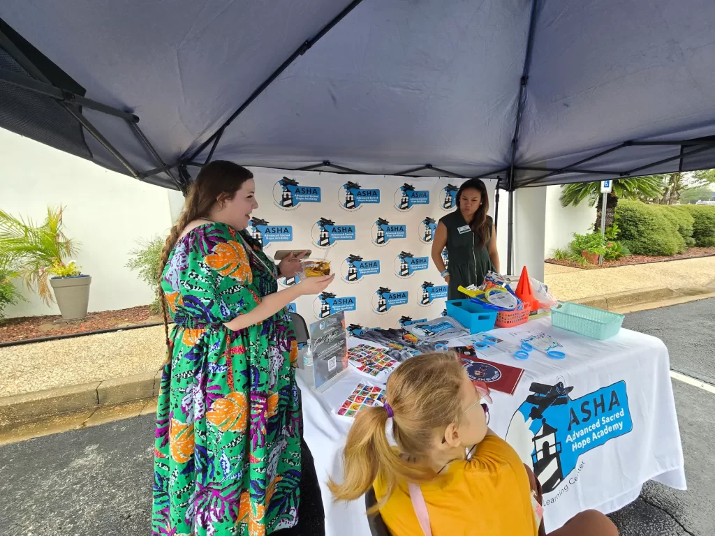 A woman in a colorful dress and another woman stand at a booth under a canopy, displaying items with the ASHA logo. A child in a yellow top is seated nearby. The booth is set up outdoors with plants and a building in the background.