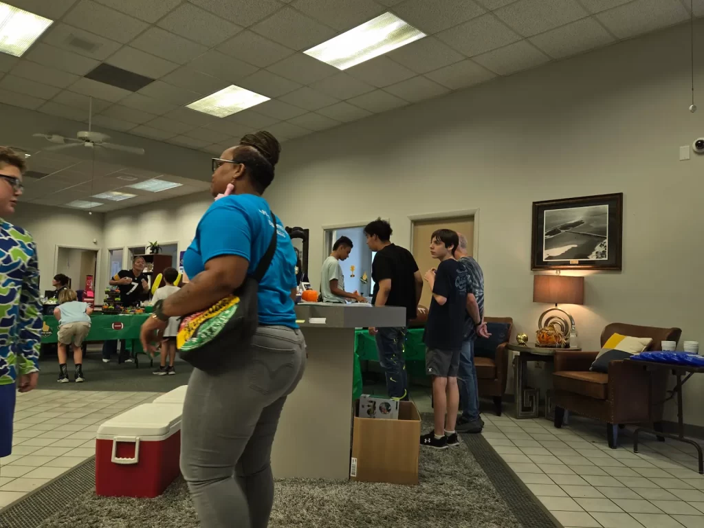 People are gathered in a room with games and seating areas. A woman with a purse stands in the foreground near a foosball table. The environment appears casual, with some individuals interacting around the tables.