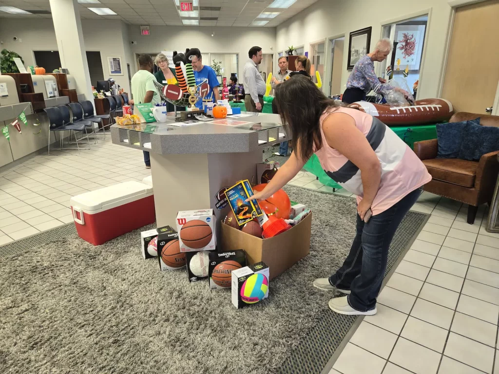 A woman bends over a box filled with various sports balls in a busy community center. People in the background interact, and the room is decorated with sports and event paraphernalia.