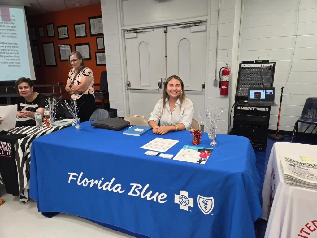 A woman sits at a booth with a blue tablecloth displaying the "Florida Blue" logo. The table has brochures and decorations. Two other women are near the booth in a room with white walls and a projector screen.