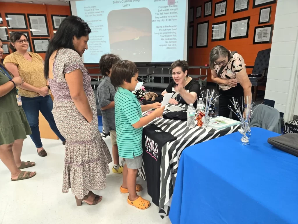 A woman is seated at a table signing items for a group of adults and children. The backdrop features a large screen with text. The table is decorated with striped fabric and shiny decorations. People are engaged and smiling.